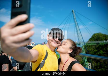 A couple shares a sweet moment taking a selfie, with one giving a kiss on the cheek, on the Langkawi Sky Bridge, Malaysia Stock Photo