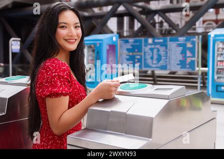 A happy woman in a red dress is using a ticket to access the public transportation system, with turnstiles in the background Stock Photo