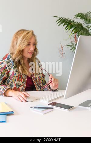 A focused blonde woman in a patterned jacket works diligently at her desk, using a computer in a modern office setting Stock Photo