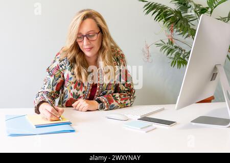 A blonde businesswoman in stylish attire focuses on writing notes at her white office desk with a computer and smartphone nearby Stock Photo