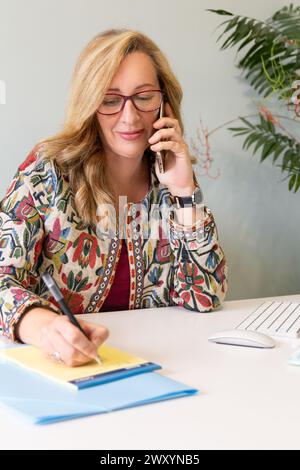 A professional blonde businesswoman is talking on the phone while taking notes in a modern office environment, showcasing multitasking and efficiency Stock Photo