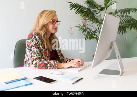 Blond businesswoman in glasses focused on her work, typing on a desktop computer with a plant beside her Stock Photo