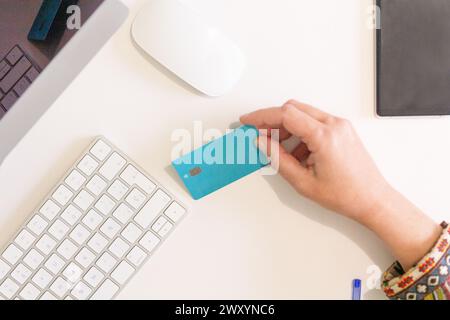 A close-up view of anonymous hand holding a teal credit card near a white keyboard, mouse, and closed tablet Stock Photo