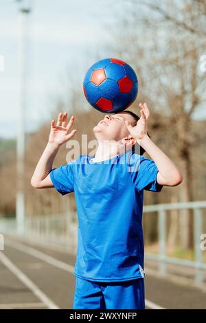 A young boy in a blue sports uniform balances a red and blue soccer ball on his head with a determined expression and clear sky in the background Stock Photo