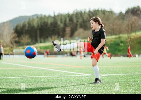 A young girl in a black shirt and red shorts kicks a soccer ball during a game on a sunny outdoor field Stock Photo