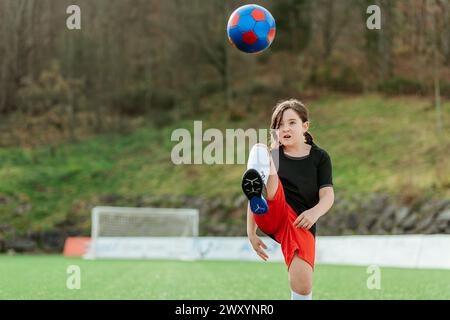 An enthusiastic young girl in soccer attire kicks a colorful ball on a grassy field with determination, showcasing youth sports Stock Photo