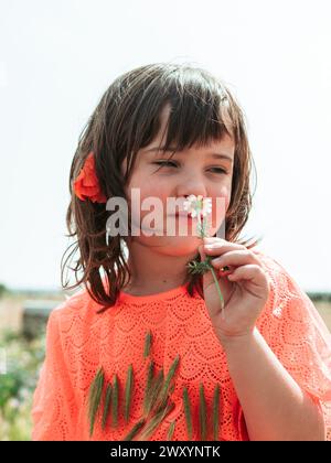 A thoughtful young girl in an orange dress examines a daisy closely, captured in a serene moment at a farm school in summer Stock Photo