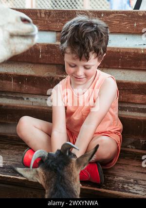 Smiling boy in summer attire enjoys a moment with a goat kid while seated on wooden steps at a farm school Stock Photo