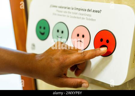 A person's finger is pressing a red unhappy face button on a customer satisfaction panel to rate the cleanliness of an airport restroom Stock Photo