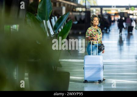 Joyful traveler with a suitcase strolling through an airport terminal, green foliage blurred in the foreground Stock Photo