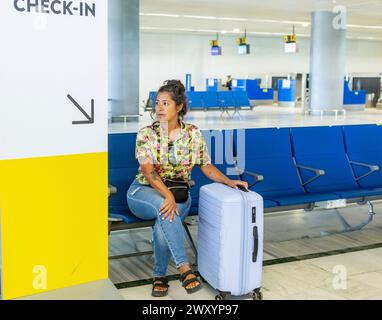A contemplative woman sits by her suitcase in the check-in area of an airport, with empty seats in the background Stock Photo