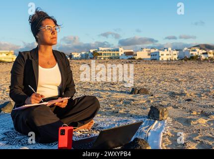 A pensive digital nomad takes notes while working from a sandy beach, with a laptop and smartphone, during a beautiful sunset backdrop. Stock Photo