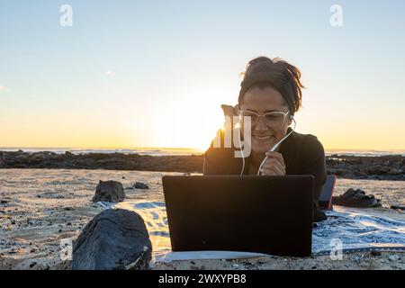 Smiling digital nomad works on a video call on laptop by the sea during a stunning sunset, embodying remote work freedom. Stock Photo