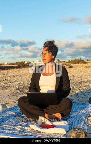 A thoughtful digital nomad sits on a beach towel, working on a laptop during sunset with a serene expression. Stock Photo