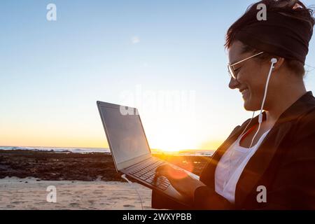 Side view of smiling woman works on her laptop on a beach at sunset, embodying the digital nomad lifestyle with headphones on and the glow of the sett Stock Photo