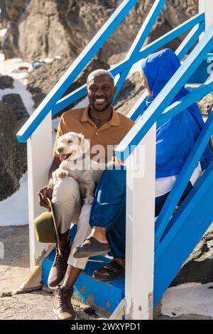 A cheerful multiethnic couple enjoys a sunny day with their happy dog on vivid blue and white stairs Stock Photo