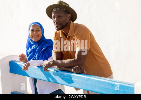 Cheerful multiethnic couple leaning on a blue railing, sharing a joyous moment under the bright sun Stock Photo