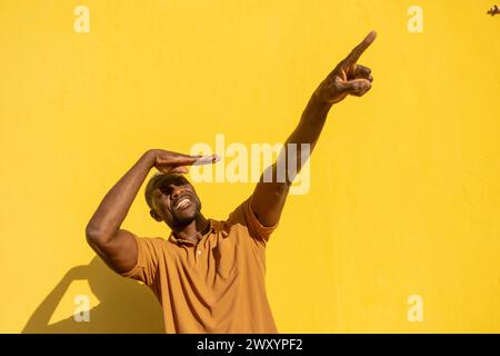 A cheerful man in a brown shirt shields his eyes and points upwards against a vivid yellow wall, expressing positivity and enthusiasm Stock Photo