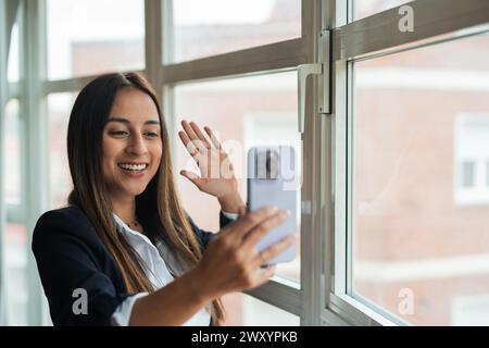 A jovial young woman in business attire takes a selfie with her smartphone, waving at the camera near a bright office window Stock Photo