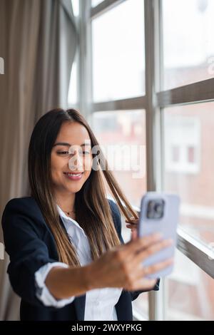 A professional woman in a suit captures a selfie with her smartphone, standing beside a window Stock Photo