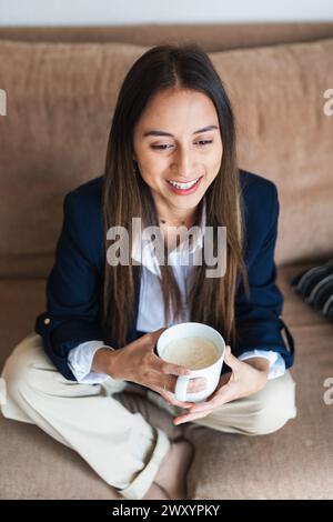 A smiling young female professional holding a cup of coffee while sitting comfortably on a sofa, taking a relaxing break Stock Photo