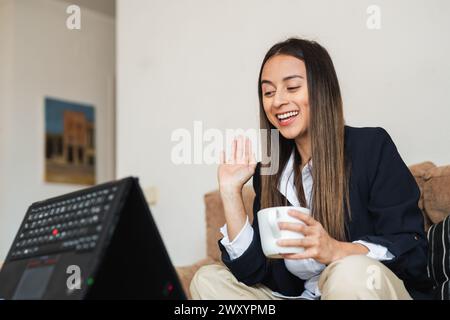 A cheerful young woman in business attire waves to a colleague over a video conference call from home, holding a coffee mug Stock Photo