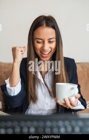 An elated businesswoman clenches her fist in victory, holding a coffee cup, while working at her desk Stock Photo