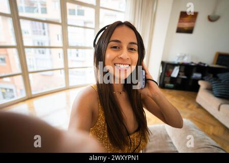 A radiant woman smiles for a selfie, her smartphone capturing the moment in a bright, cozy living room setting Stock Photo