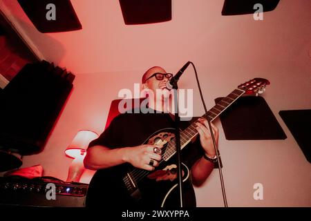 From below male guitarist sings passionately with eyes closed into a microphone, strumming his guitar in a dimly lit room Stock Photo
