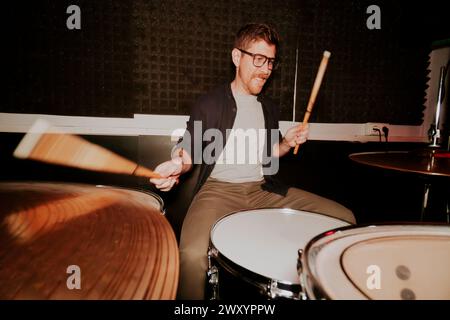 Energetic male drummer with glasses in motion, playing drums with a smile, in a room with dark ambiance, not looking at the camera. Stock Photo