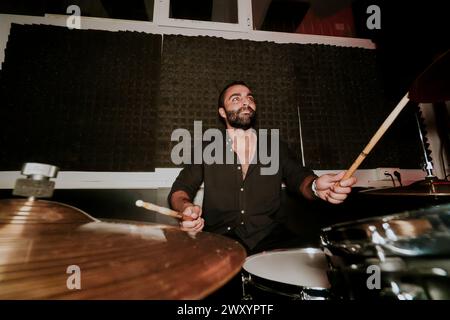 A bearded man ardently plays the drums, sticks in hand, looking away from the camera in a dimly lit studio setting, exuding a vibe of intense focus an Stock Photo