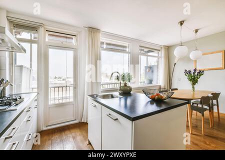 A spacious and well-lit modern kitchen featuring white cabinetry, hardwood floors, a central island, dining table setup, and open balcony doors inviti Stock Photo
