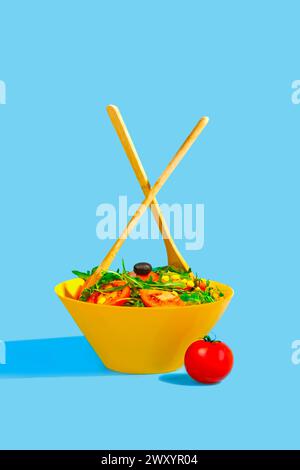 A vibrant fresh salad served in a yellow bowl with two wooden spoons crossed over it, isolated on a blue background with a single tomato aside Stock Photo