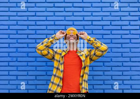 Cheerful man in a yellow hat adjusts his orange sunglasses, expressing joy against a textured blue brick wall backdrop Stock Photo