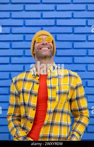 A man radiates happiness with a beaming smile, sporting a yellow beanie and sunglasses against a bright blue brick wall Stock Photo