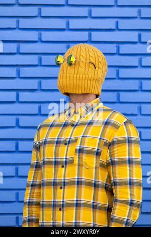 Anonymous man in a yellow beanie covering his face stands against a blue brick wall, creating an air of mystery and humor Stock Photo