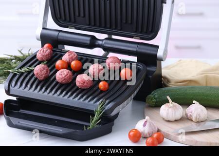 Electric grill with meatballs, tomatoes and vegetables on white table in kitchen Stock Photo