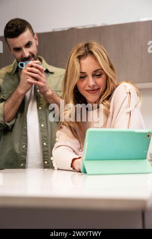 Smiling happy man drinking coffee and woman using a touch pad in a video call. Lovely handsome couple looking tablet together in the kitchen. Stock Photo