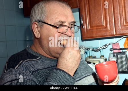 The picture shows an elderly man, he is eating a bun and holding a cup in his hand. Stock Photo