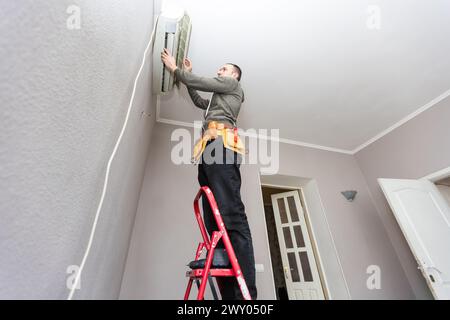 Professional electrician man with screwdriver maintaining, cleaning modern air conditioner indoors. Young technician standing on a ladder in the room Stock Photo