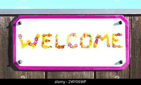 WELCOME from yellow pink red and orange flowers collage, enamel, metal, elongated stone pottery sign on a rustic wooden wall. pink frame, welcome card Stock Photo