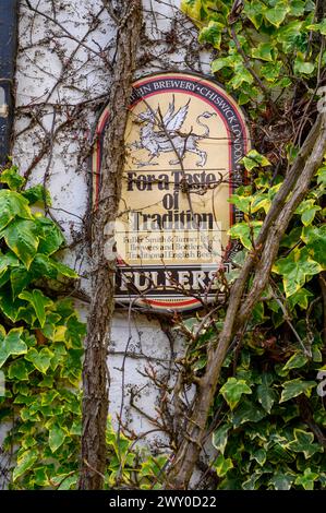 Vintage enamel sign for Fullers beers partly covered by vines on the wall of The Rising Sun pub in Nutbourne village in West Sussex, England. Stock Photo