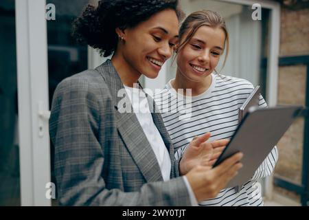 Two busy female coworkers discussing project standing at office terrace and use digital tablet Stock Photo