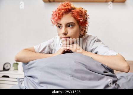 relaxing red haired queer person in homewear sitting on her bed at home and looking away, leisure Stock Photo