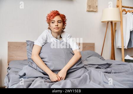 relaxing red haired queer person in homewear sitting on her bed at home and looking away, leisure Stock Photo