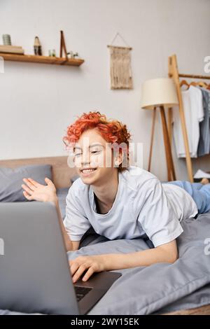 good looking red haired cheerful queer person in homewear having video call while lying in bed Stock Photo