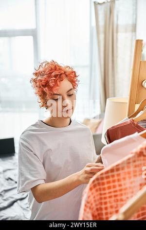 beautiful red haired extravagant person picking up stylish clothes near rack at home, leisure time Stock Photo
