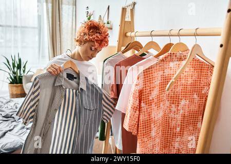 beautiful red haired extravagant person picking up stylish clothes near rack at home, leisure time Stock Photo
