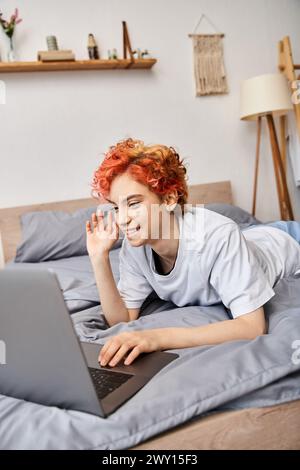 good looking red haired cheerful queer person in homewear having video call while lying in bed Stock Photo