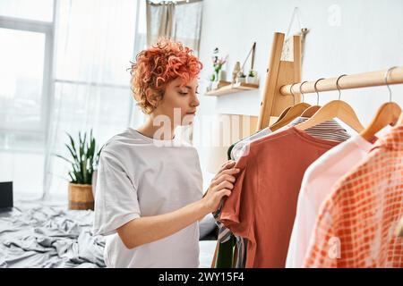 attractive red haired extravagant person picking up stylish clothes near rack at home, leisure time Stock Photo
